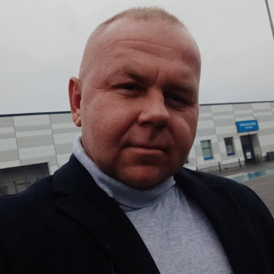 Man with a shaved head wearing a grey turtleneck and dark jacket outside a building.